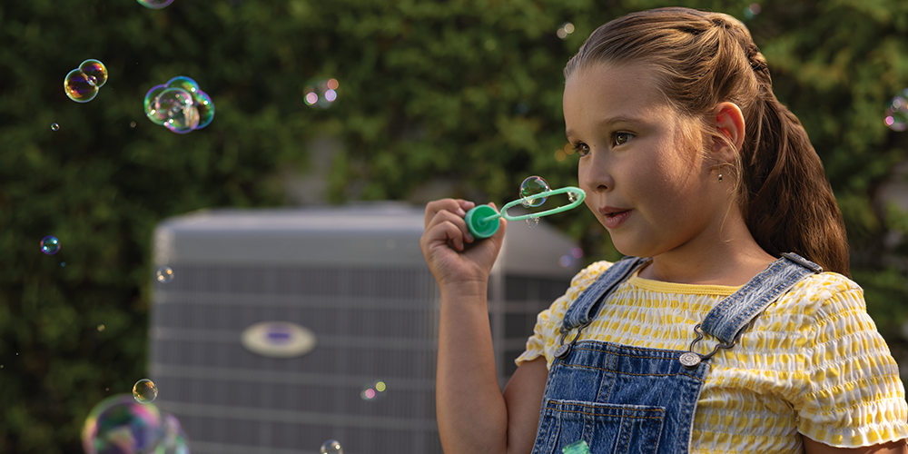 Little girl in overalls blows bubbles in front of air conditioner in background.