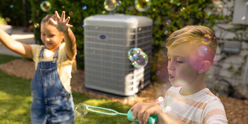 Young brother and sister play with bubbles in backyard in front of new air conditioner.